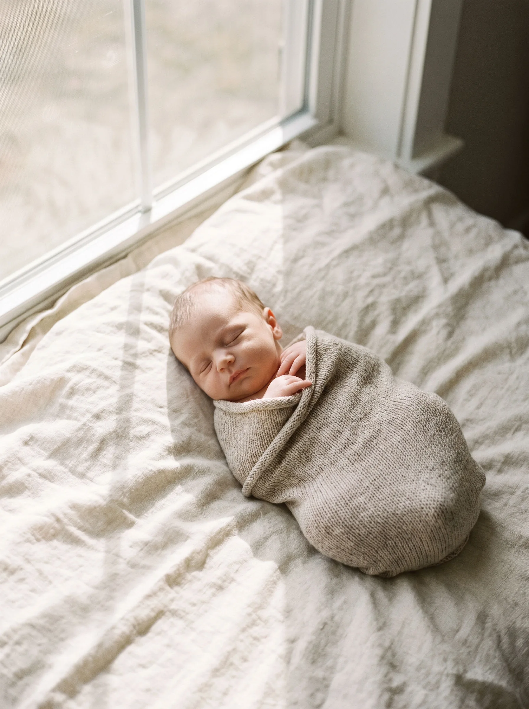 A newborn asleep in a knit wrap on a linen blanket by the window
